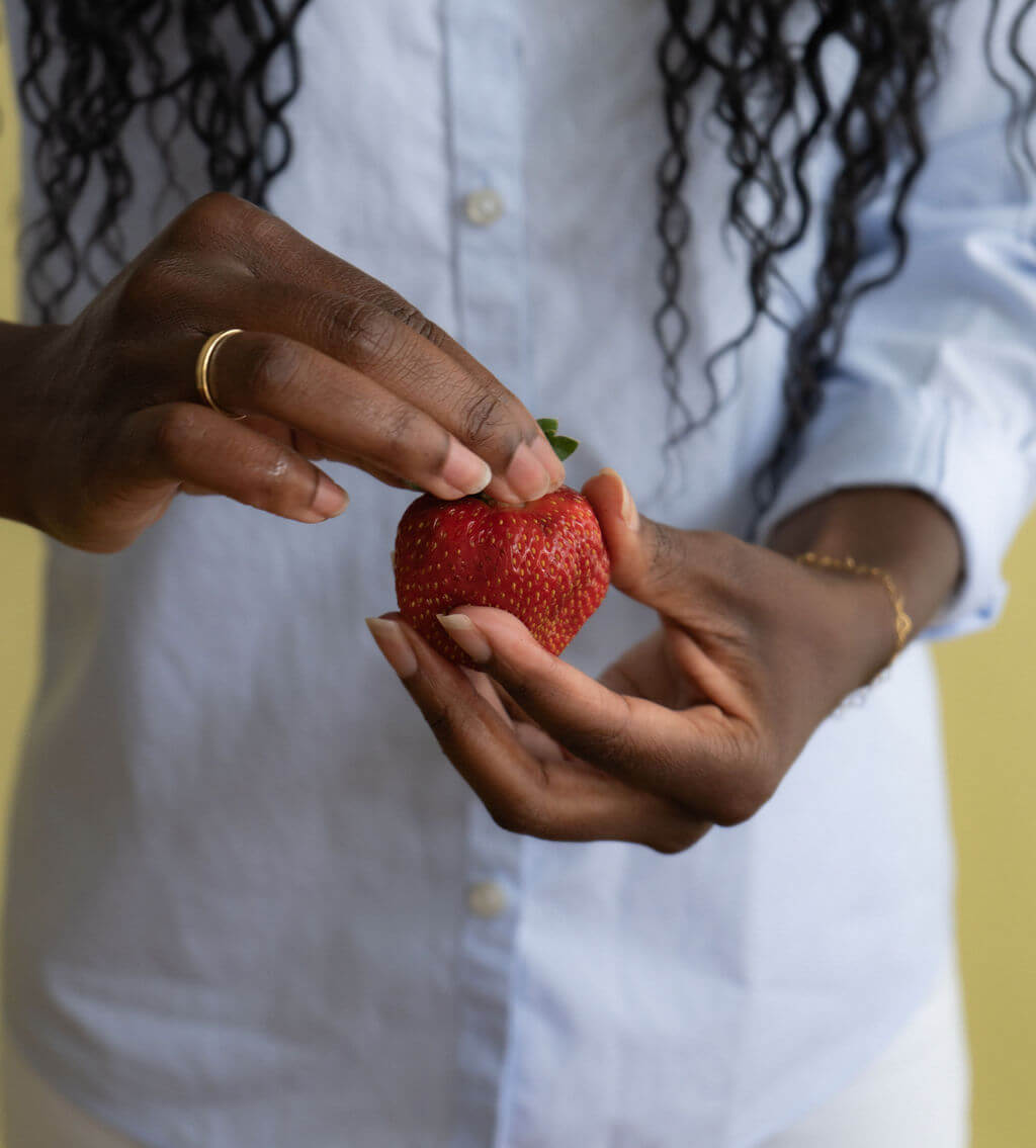 Close-up of hands holding a ripe red strawberry against a white shirt, highlighting its seeds and fresh green leaves in a calm moment.
