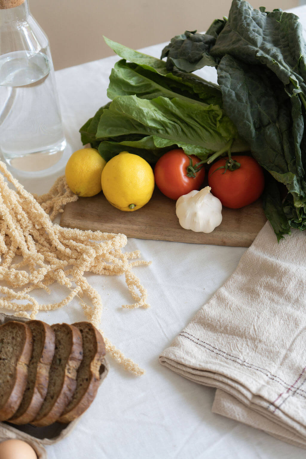 Fresh lettuce, kale, tomatoes, lemons, and garlic on a wooden board beside sliced bread, a glass bottle, and linen cloths; a rustic, wholesome healthy kitchen scene.